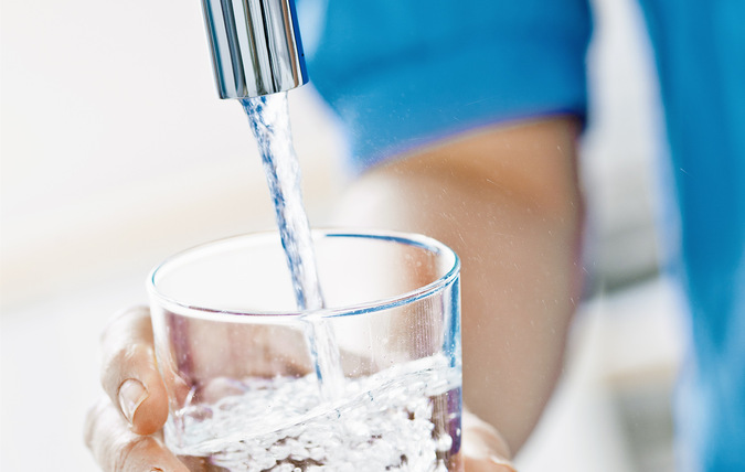 Ett glas som fylls upp med vatten. En hand håller glaset under en vattenkran. Hen har blå tröja på sig.
