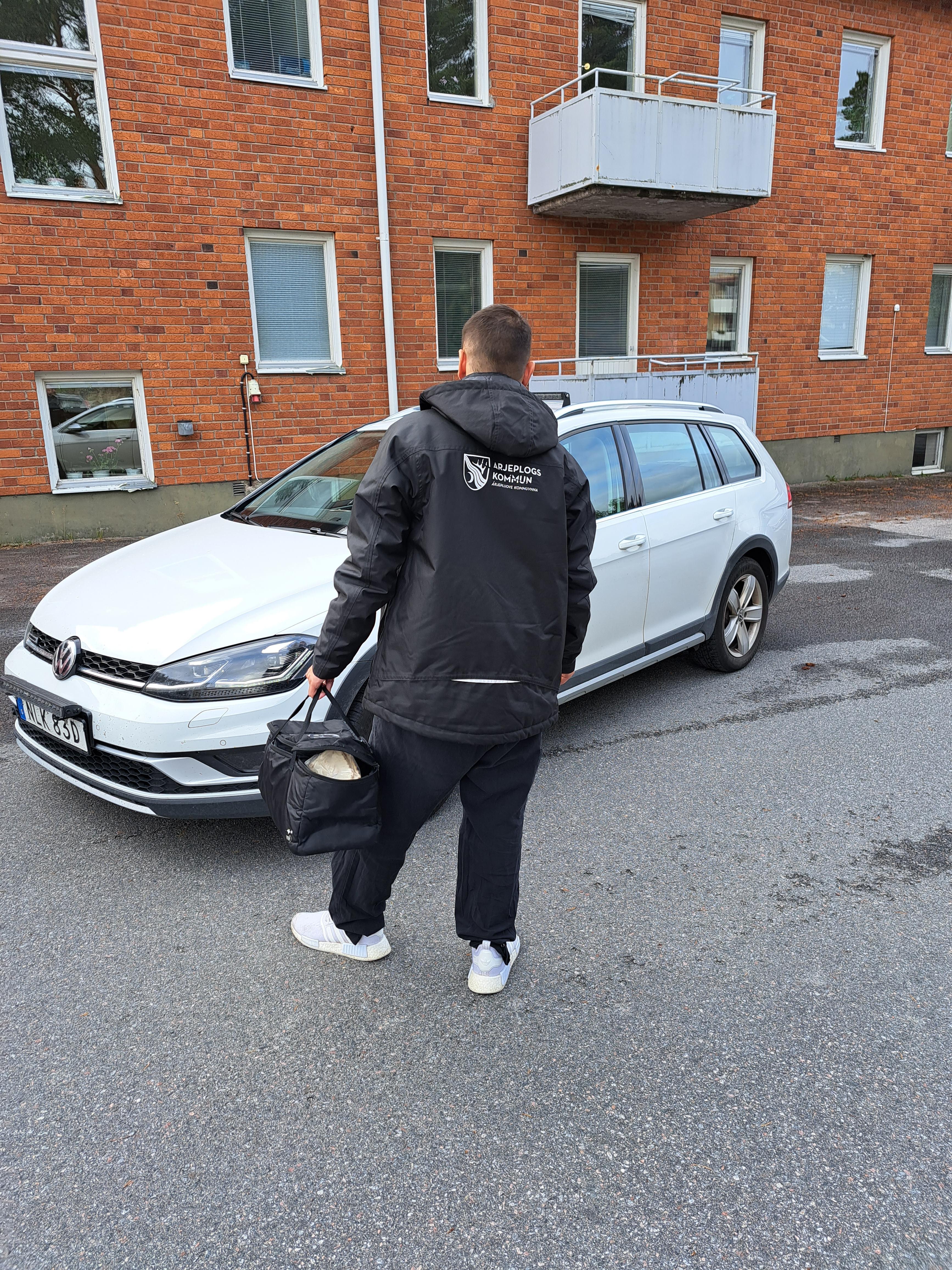 En man i en svart jacka med texten Arjeplogs kommun på ryggen står framför en bil, med en väska i handen.