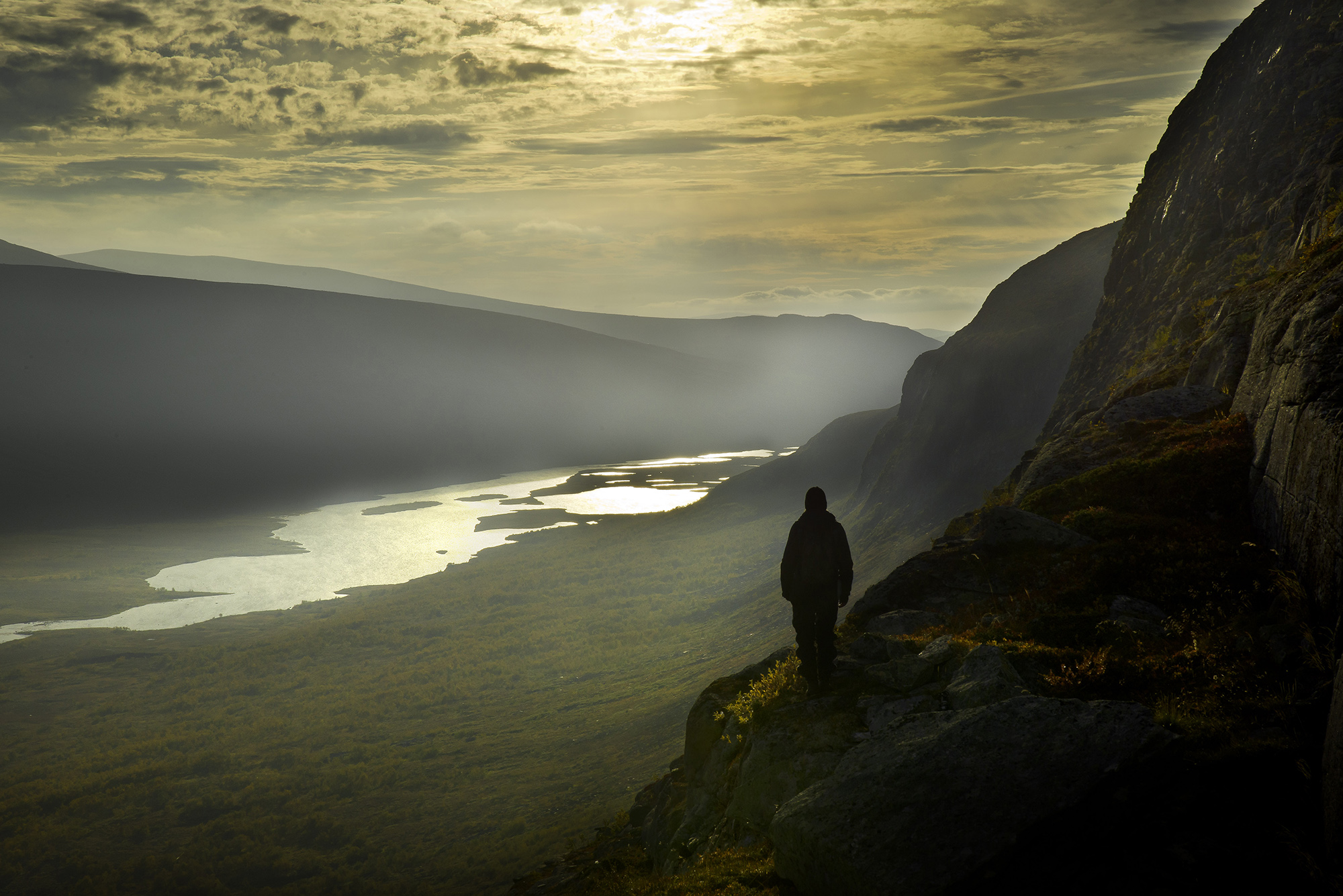 En man står på ett berg och blickar ut över en dal med en sjö.
