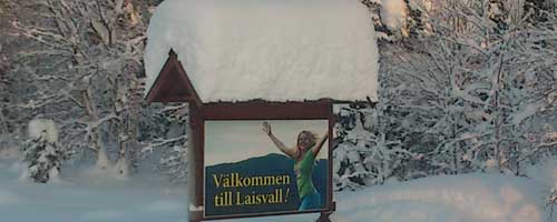 En skylt det står "Välkommen till Laisvall" på, där en tjej håller upp händerna i en glad gest.